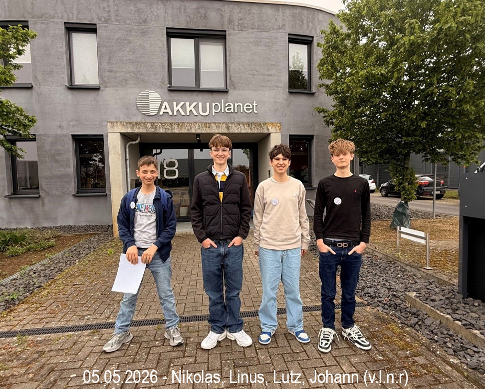 Vier junge Männer lächeln vor einem modernen, grauen Firmengebäude mit Schriftzug und Logo „AKKUplanet“. Von links: Nikolas mit Blatt Papier; Linus in dunkler Steppjacke; Lutz und Johann mit Ansteckern. Gepflasterter Boden.