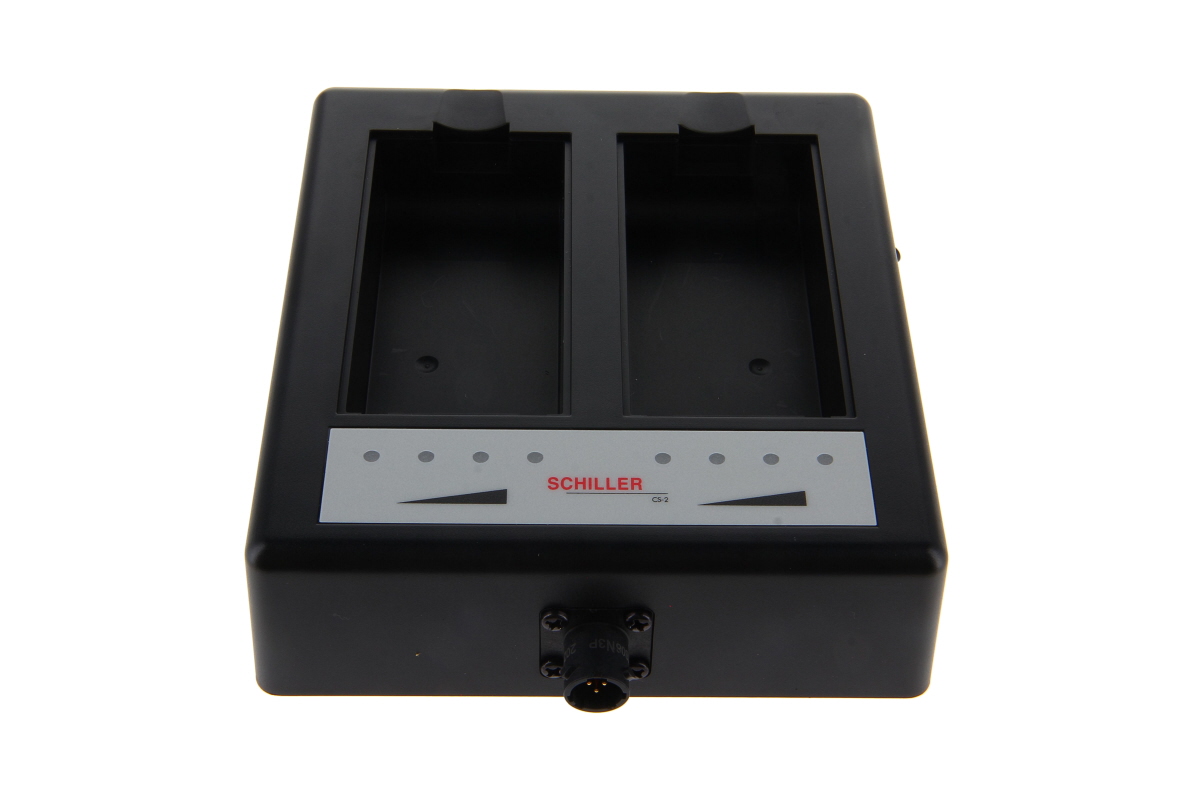 Black ‘ Schiller ’ CS-2 charger with two vertical charging slots for Li-ion batteries. Below this is a light grey control panel with red ‘Schiller CS-2’ lettering, four status LEDs and a triangular charge indicator. Round connector at the front.